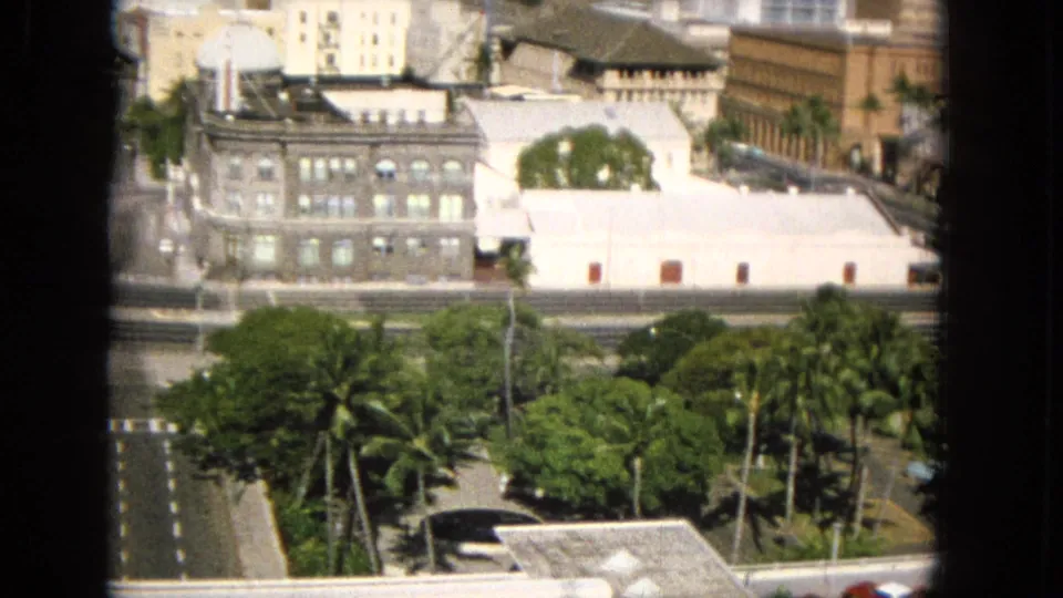 1965 8mm frame likely showing Honolulu from an elevated interior or balcony vantage, with palms, a multi-lane road, low civic-looking buildings, and dark window mullions in front.