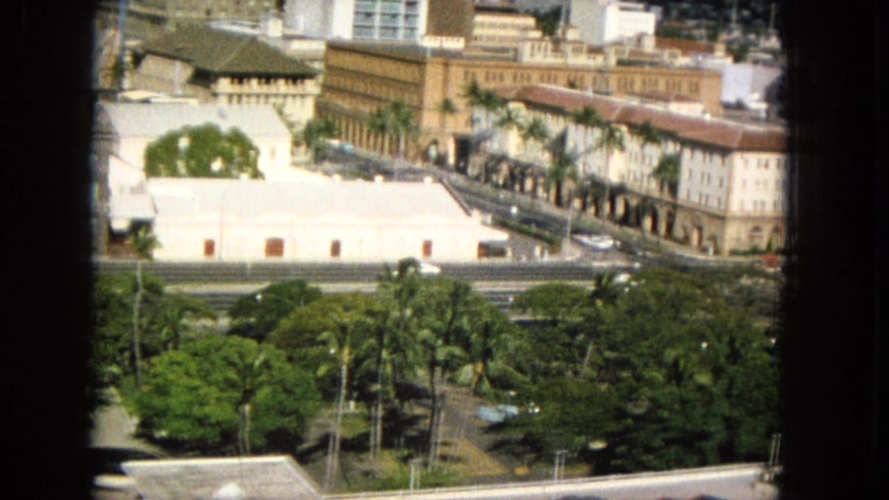 Palm-lined urban view with an arched tan building, roadway, high-rises, and distant hillside.
