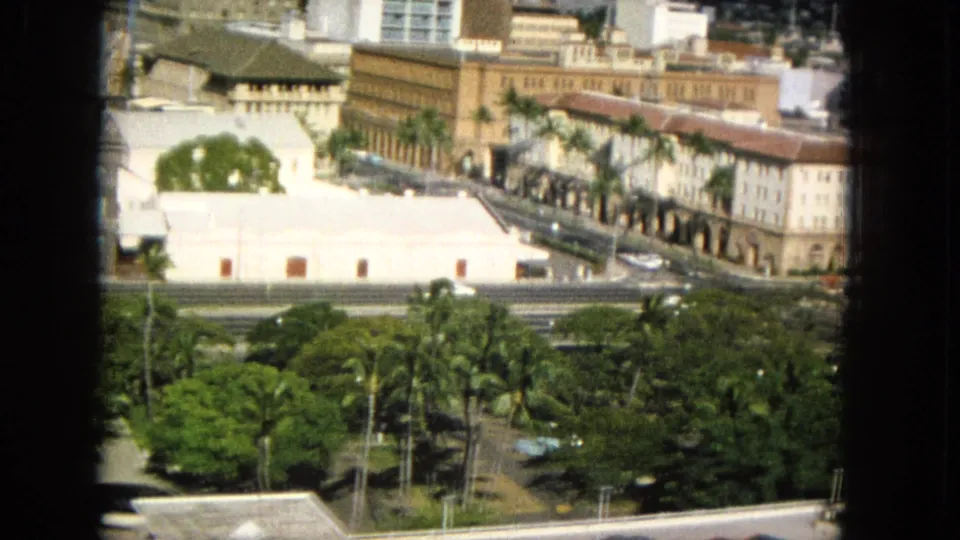 1965 8mm frame likely showing Honolulu architecture and traffic, including a long tan building with arched colonnades, palms, roadways, high-rises, and a hillside beyond.