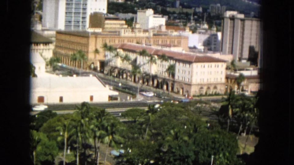1965 8mm frame likely showing a Hawaiian downtown district with foreground greenery, a pale low building, a long arched facade, cars on city streets, and taller buildings behind.