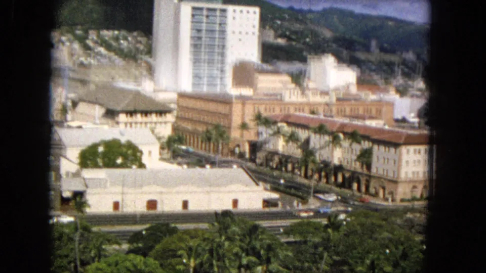 1965 8mm frame likely showing Honolulu through dark window framing, with palm trees, a road carrying cars, low civic-looking buildings, high-rises, and hillside terrain in the distance.