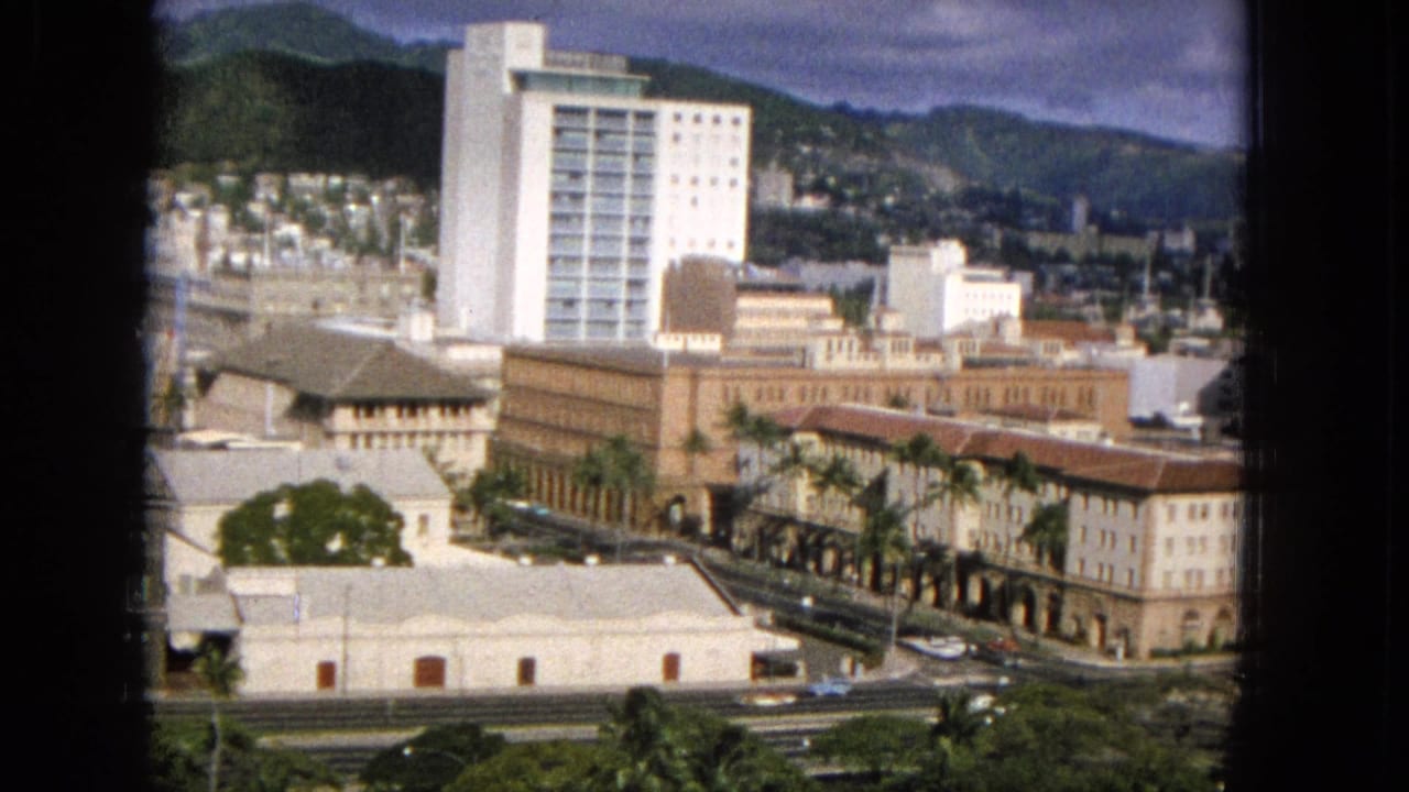 Elevated 1960s Hawaii city view, possibly Honolulu, showing a white high-rise tower, older brown urban buildings, palm trees, cars, and green mountains in the distance.