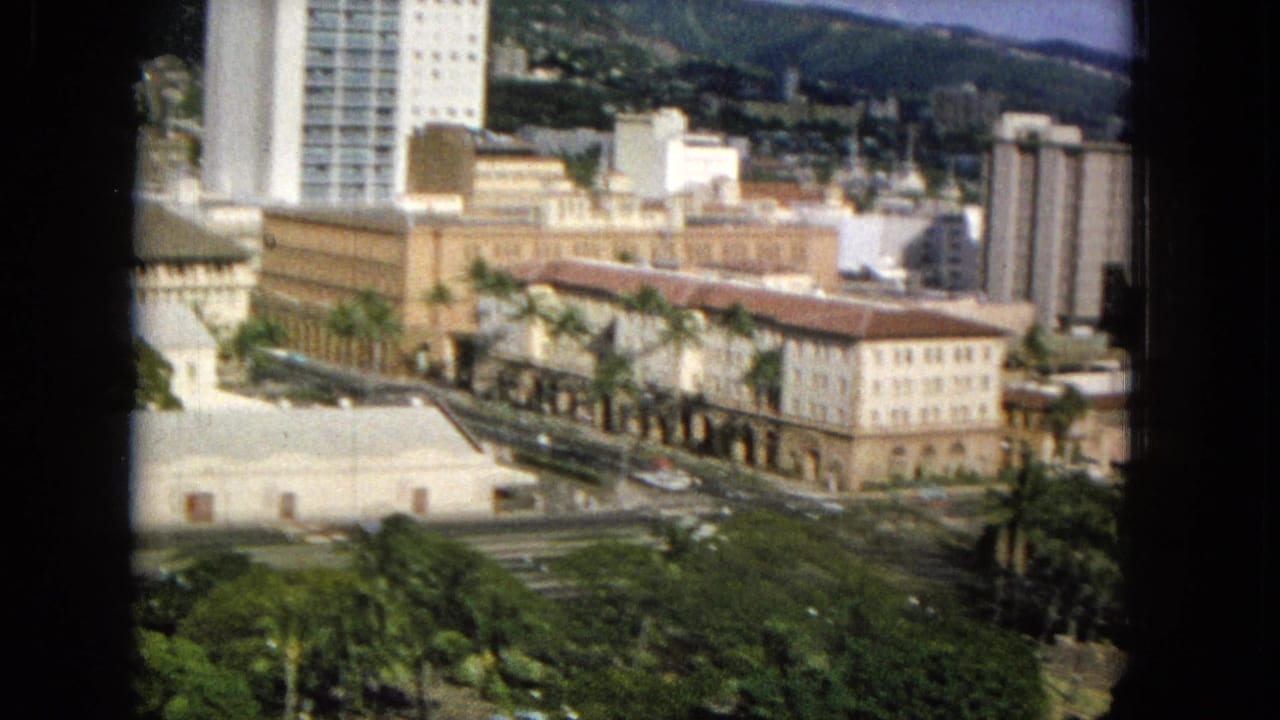 City blocks with palm trees, a tall white building, older masonry buildings, roads, cars, and hillside background.