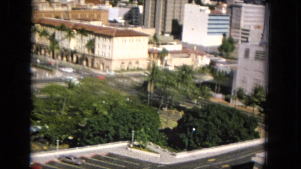 Closer elevated view of a Hawaiian urban block in 1960s footage, with cream and brown buildings, palms, street traffic, dense greenery, and a bright foreground wall.