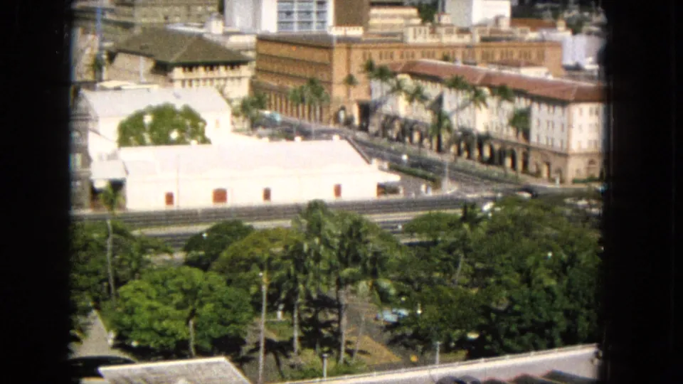 Elevated 1960s Hawaii city view showing a bright white foreground wall, palms, long urban buildings, roadways with cars, leafy trees, and taller buildings beyond.