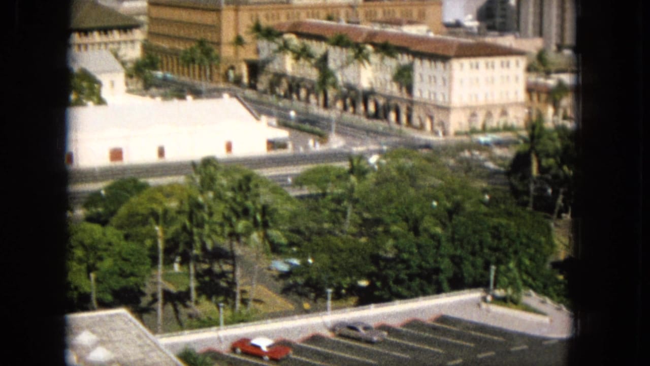 1965 Super 8 frame from Hawaii showing an elevated view through a dark window frame toward palm-lined grounds, a street, arched architecture, and classic parked cars.
