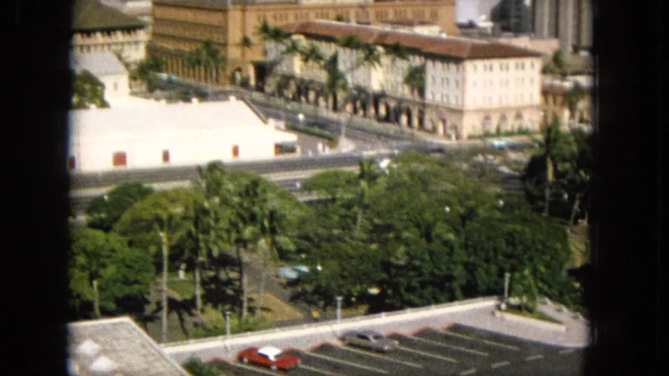 1965 Super 8 frame from Hawaii showing an elevated view through a dark window frame toward palm-lined grounds, a street, arched architecture, and classic parked cars.