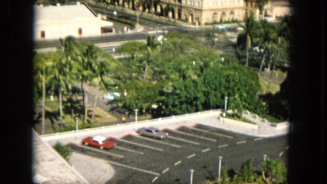 1965 Super 8 frame from Hawaii with a window-framed elevated view of a parking lot, red and silver cars, dense trees, a roadway, and a pale arched building.