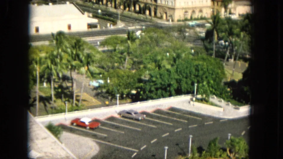 1965 Super 8 frame from Hawaii with a window-framed elevated view of a parking lot, red and silver cars, dense trees, a roadway, and a pale arched building.