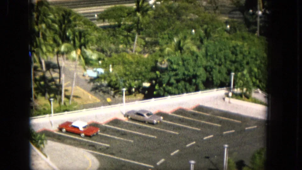 1965 Super 8 frame from Hawaii showing a closer elevated view of green landscaping, street markings, a parking lot, and classic cars including a red car and silver sedan.