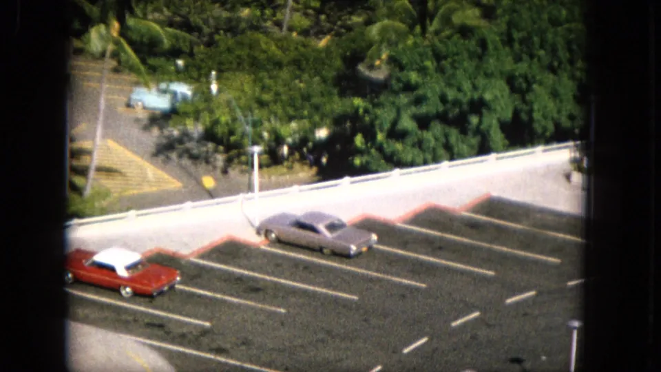 1965 Super 8 frame from Hawaii showing a parking lot with red and silver cars, a low wall, light poles, dense tropical trees, and a dark window edge in the foreground.