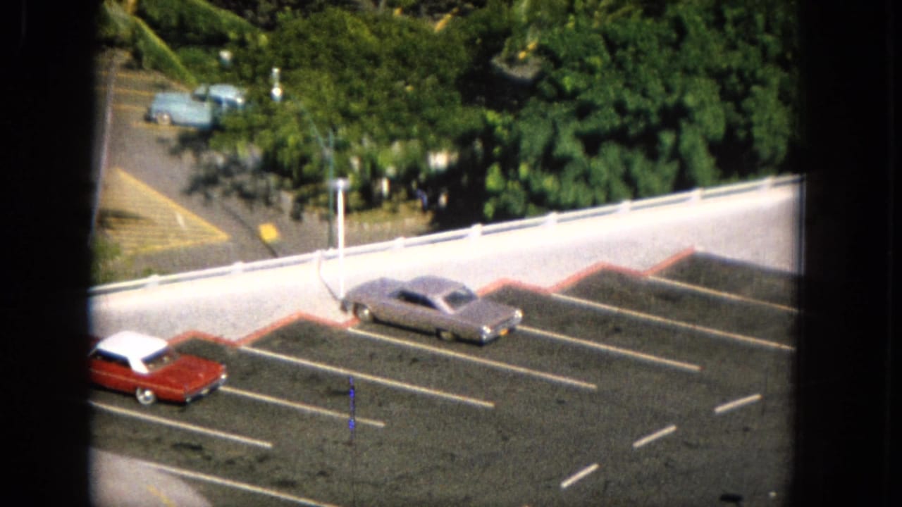 1965 Molokai home-movie frame showing a gray sedan and red car parked in marked asphalt spaces below tropical greenery and a white roadside railing.