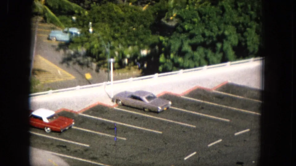 1965 Molokai home-movie frame showing a gray sedan and red car parked in marked asphalt spaces below tropical greenery and a white roadside railing.