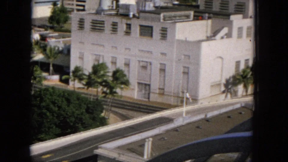 Super 8 frame from an elevated position showing a white building with palms along the street, a road below, and a blue arched structure crossing the foreground.
