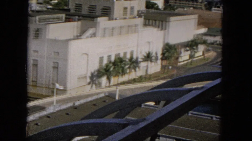 Elevated 1965 Super 8 view across a blue arched foreground structure toward a palm-lined white building, road, and sunlit tropical streetscape.