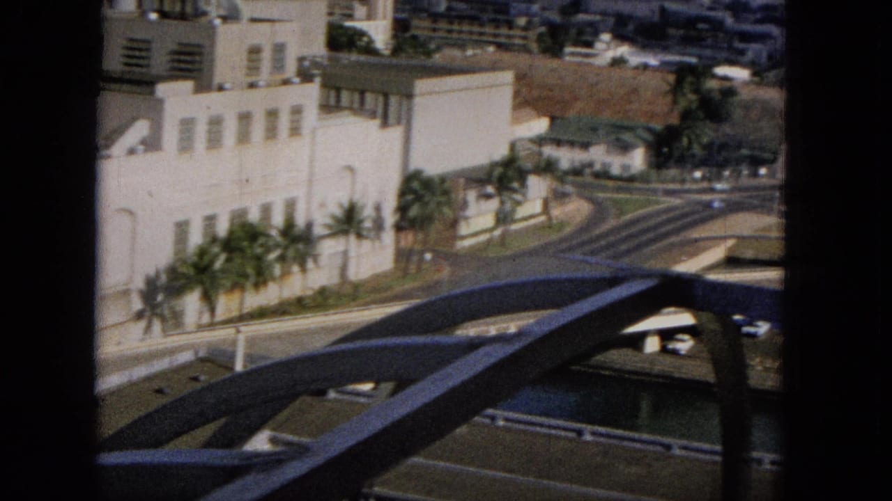 1965 8mm travel footage appears to look from an elevated position across a Hawaiian urban waterfront with a pale building, palms, roadways, and a bridge span.