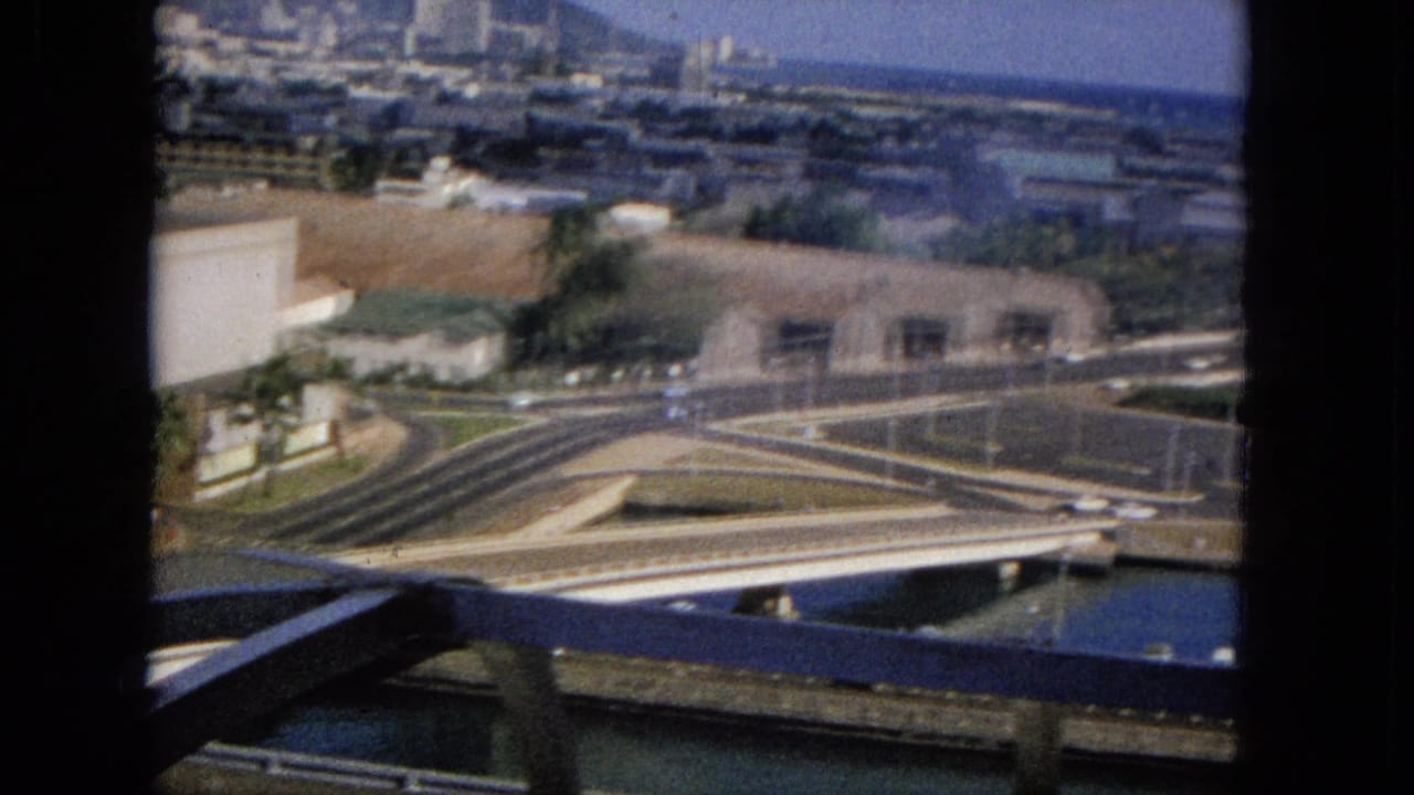 Bridge and freeway interchange seen through dark vertical bars with buildings, cars, water, and distant shoreline beyond.