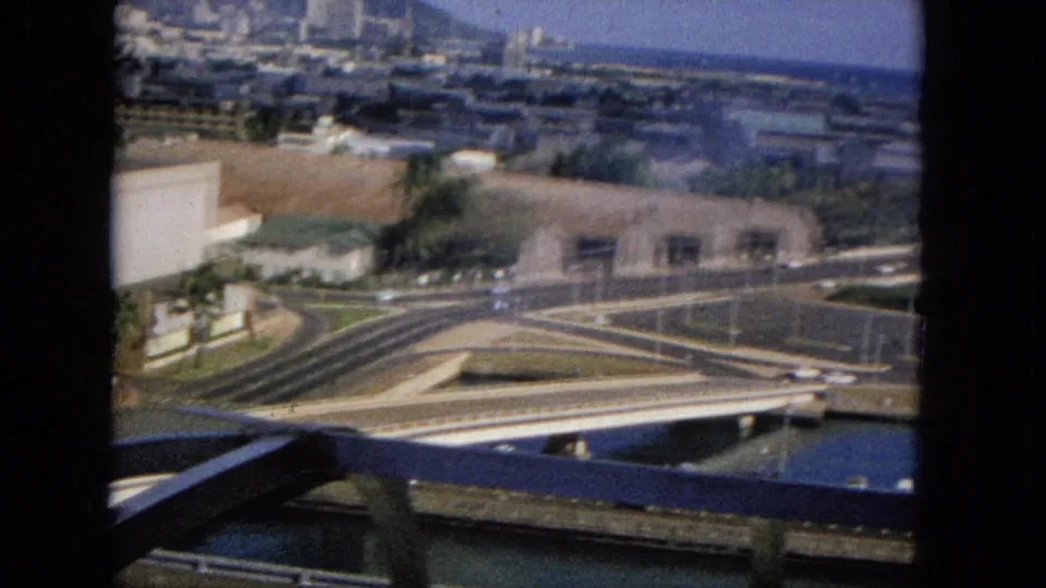 The view opens across a bridge or freeway interchange, small cars, waterfront buildings, and a distant ocean horizon in archival 1960s home-movie footage.