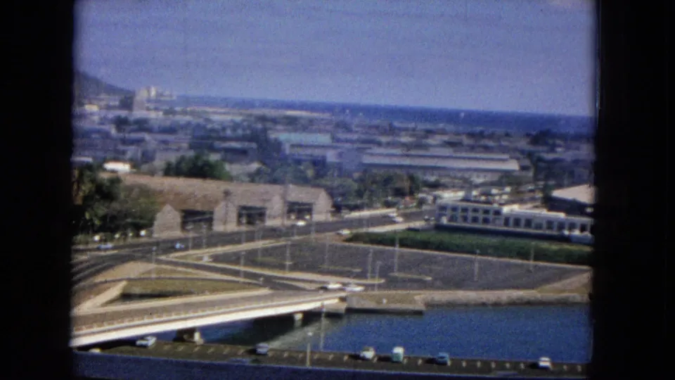 An elevated 1965-era home-movie frame shows a broad urban waterfront with bridge lanes, parked cars, low buildings, water, distant hills, and ocean beyond.