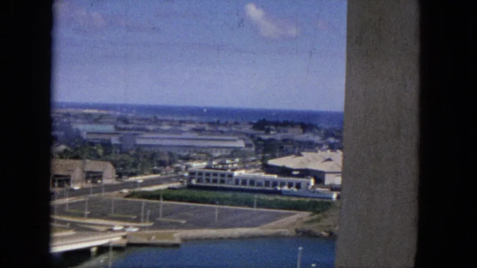 The final still continues the elevated waterfront view, with bridge structure, roads, parked cars, low buildings, open water, and a vertical wall or column at the edge of frame.