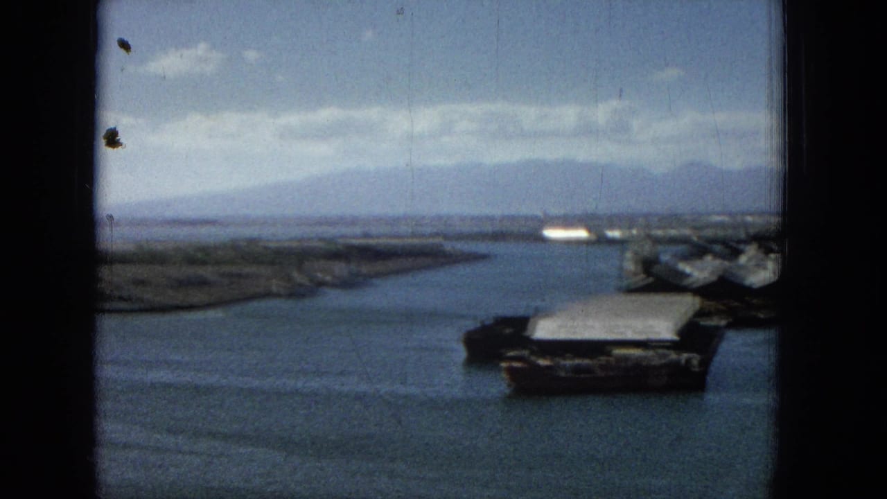 1965 Hawaii home-movie frame showing calm harbor water, a low shoreline, distant landforms, and waterfront structures through a dark window frame.
