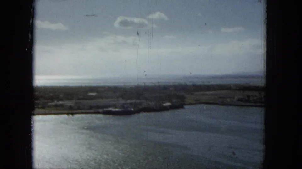Hazy 1965 Hawaii waterfront frame with calm water, low shoreline, clouded sky, and dark window framing visible around the scene.