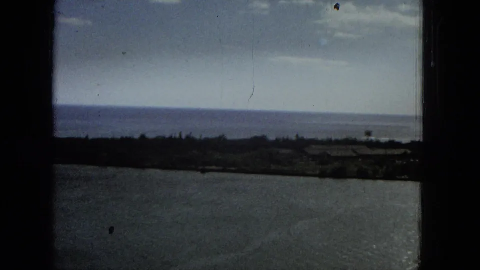 1965 Hawaii home-movie frame showing a darker coastal water view, low shoreline silhouettes, distant horizon, and visible film wear through a window frame.