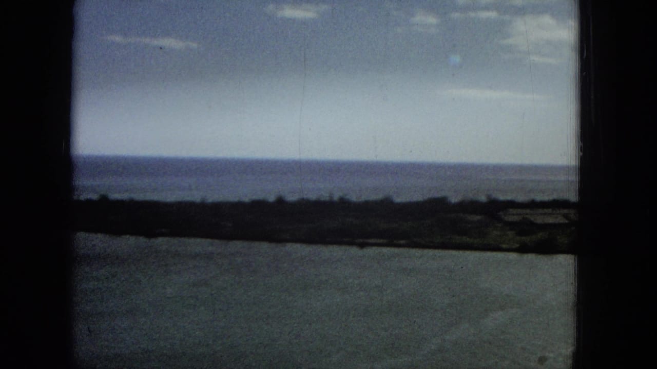 1965 Super 8 frame showing calm ocean water, a distant low coastline, pale sky, and dark vertical and horizontal foreground framing from a high vantage point.