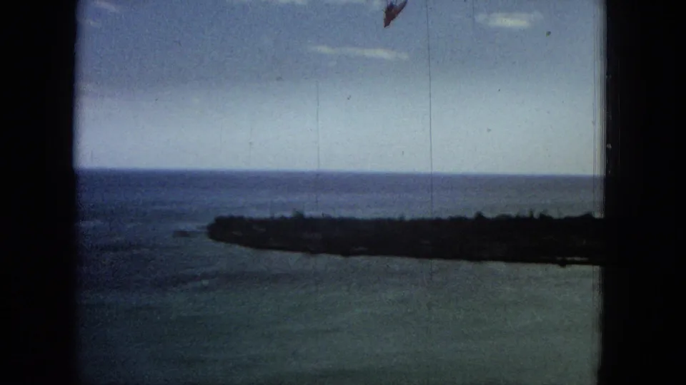 1965 Super 8 frame with blue ocean, low dark shoreline, clear horizon, and heavy dark foreground framing suggesting a view from behind a window or structure.