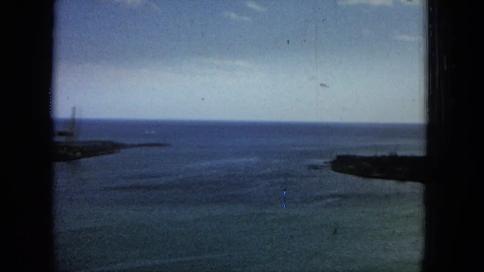 1965 Super 8 frame looking over harbor water toward low coastal land, with the view partially blocked by dark structural framing at the sides and center.