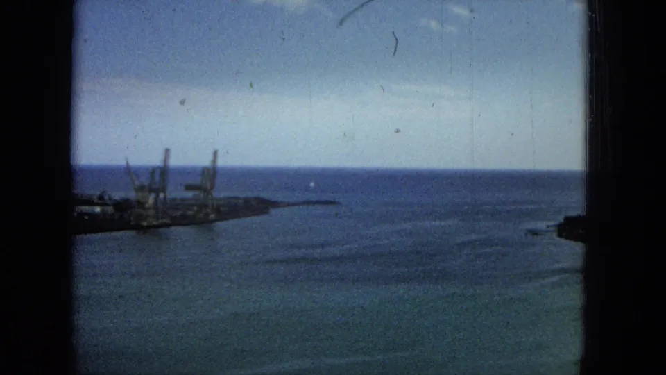 1965 Super 8 frame showing harbor infrastructure with ships or docked vessels, crane-like structures, open ocean, a low shoreline, and dark framing at the image edges.