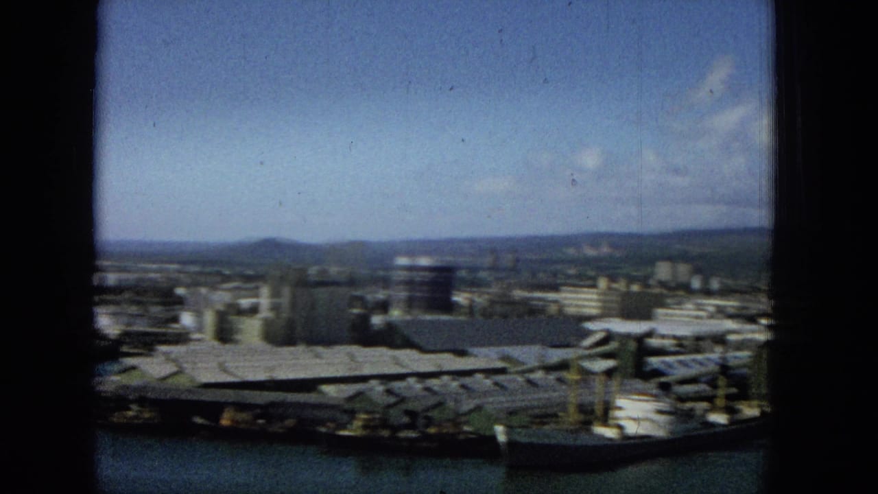 A 1965 8mm frame shows a coastal city harbor with dock roofs, boats, industrial buildings, distant hills, and blue sky viewed through a dark window frame.