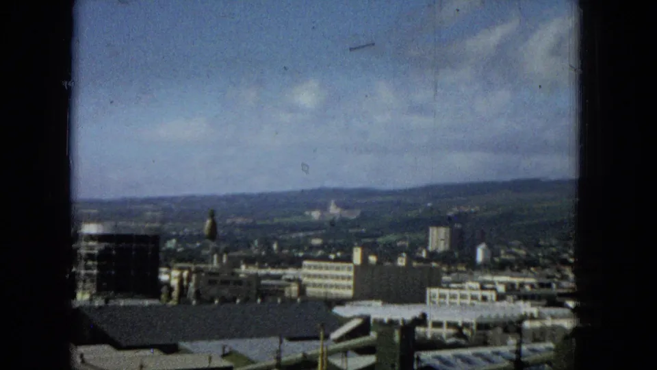 A later frame shifts slightly across the cityscape, showing a dark foreground window frame, rooftops, a tall building, clustered urban blocks, hills, and a partly cloudy sky.