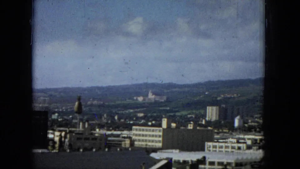 The final frame holds on the city and surrounding hills, with harbor-side roofs and mid-rise buildings below a soft blue sky marked by home-movie film artifacts.