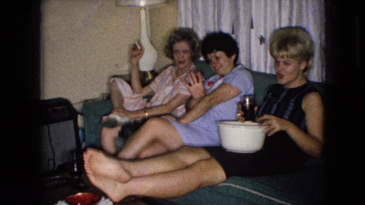 In a 1965 Wellsburg, Iowa home-movie frame, three adult women relax on a green couch in a living room, with one holding a cigarette and another near a drink and white bowl.