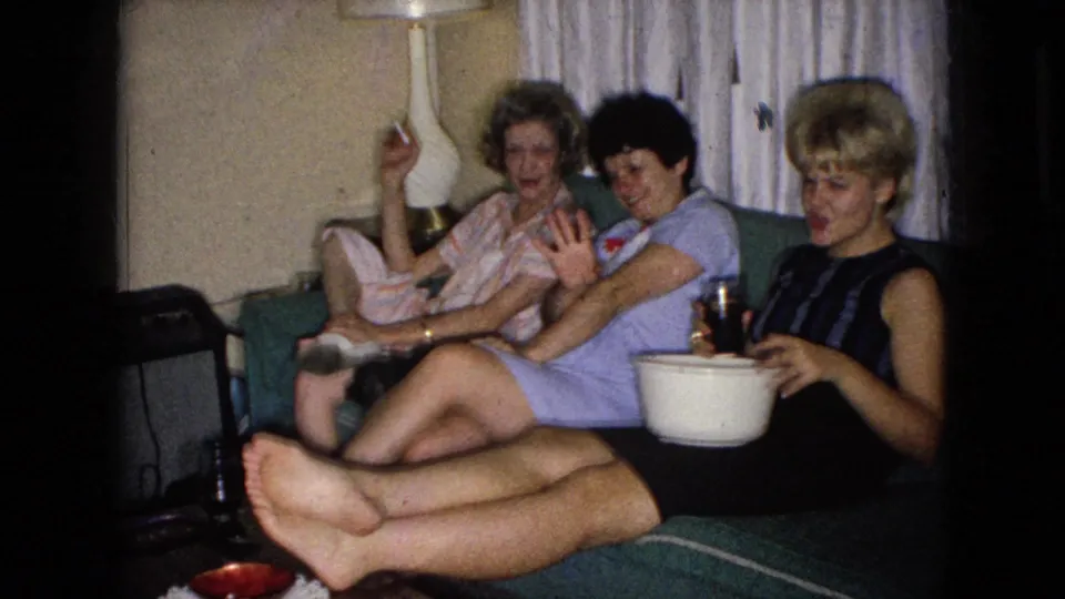 In a 1965 Wellsburg, Iowa home-movie frame, three adult women relax on a green couch in a living room, with one holding a cigarette and another near a drink and white bowl.