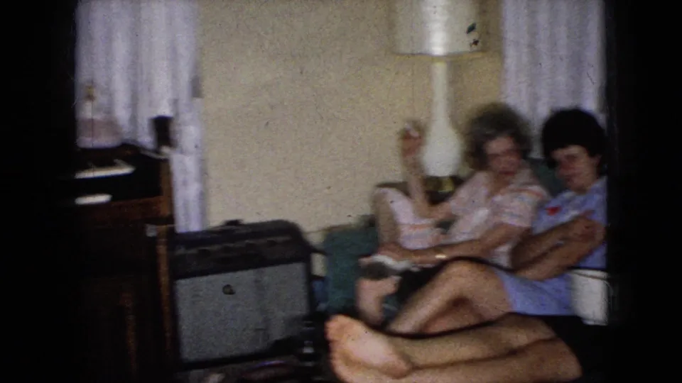 In the 1965 home-movie frame, two adult women sit close together on a green couch with a lamp, curtains, a side table, and a small television or cabinet nearby, while a third woman is partly cropped at right.