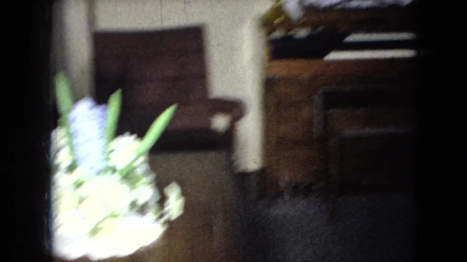 The final 1965 home-movie frame shifts away from the women to a soft, blurred living-room view with a chair, upright piano, white floor lamp, and bright flowers or plant near the camera.