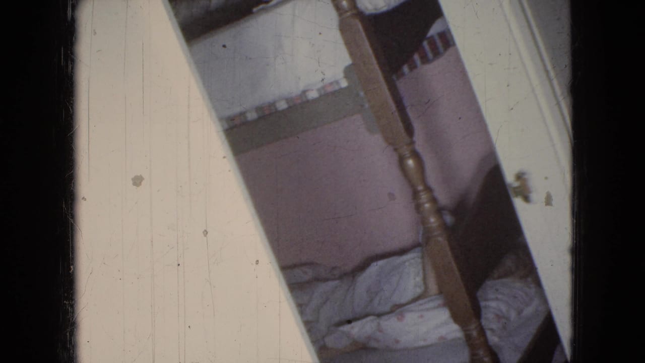 1965 home-movie frame from a private interior in Wellsburg, Iowa, showing a doorway view of a wooden bunk bed and soft bedding in a bedroom.