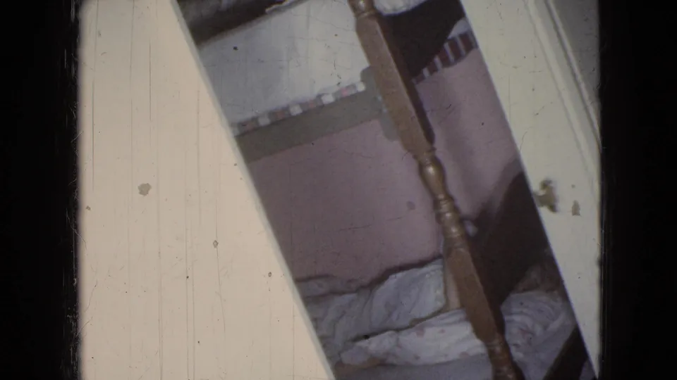 1965 home-movie frame from a private interior in Wellsburg, Iowa, showing a doorway view of a wooden bunk bed and soft bedding in a bedroom.