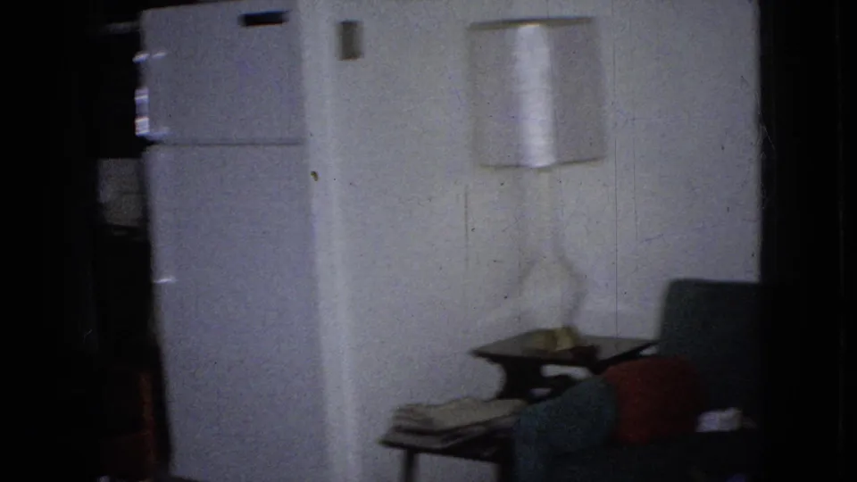 Handheld 8mm frame of a living room corner with curtains, a wall lamp, a small table, a sofa edge, and household items in a private Iowa interior.