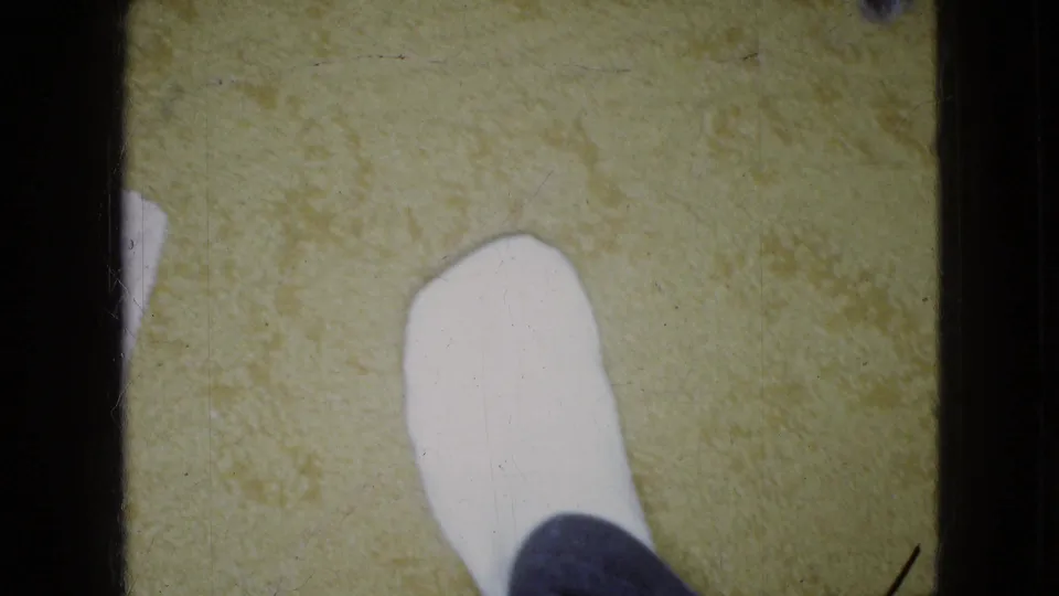 A white shoe or socked foot rests on yellow carpet while the small dog is largely outside the frame, preserving a close domestic floor view from the 1965 home movie.