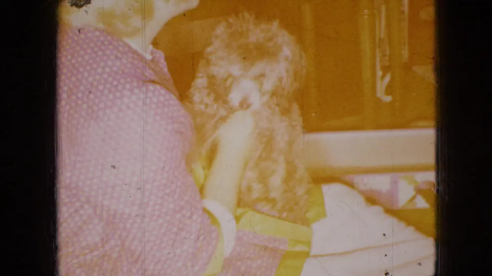 In this 1965 home-movie frame from the United States, a blonde person sits indoors holding a small light-colored dog, with warm exposure and visible film wear.