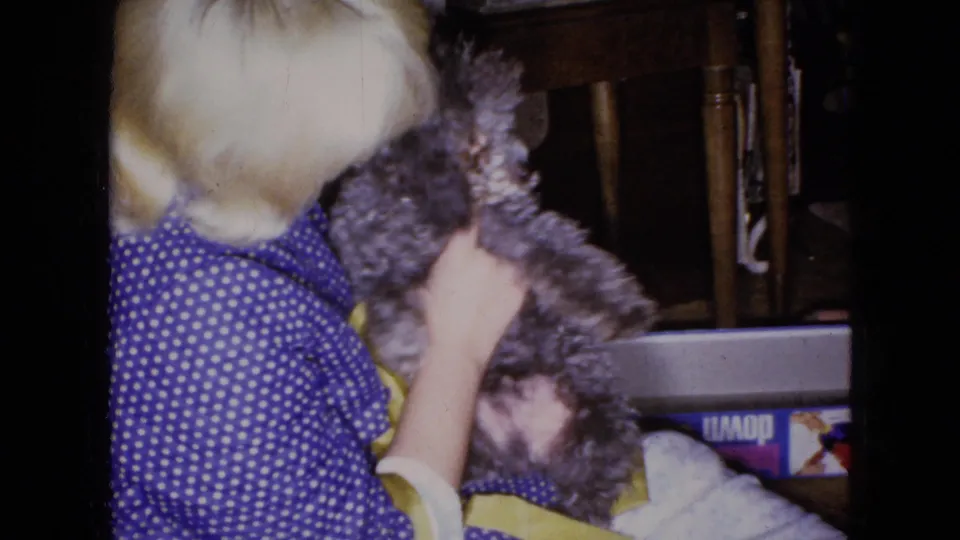 The blonde subject turns toward the small gray dog while holding it close in a private indoor setting, captured with grain and slight motion softness typical of 8mm footage.