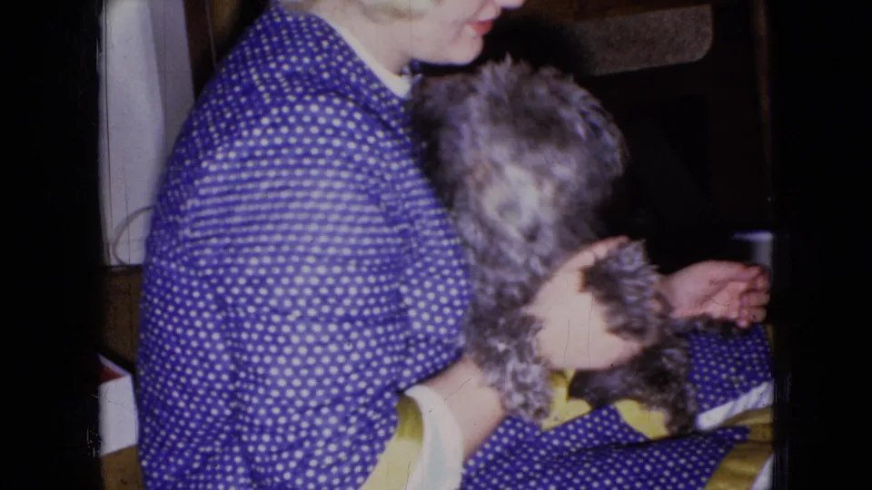 In 1965 home-movie footage from Wellsburg, Iowa, a seated woman in a blue and white polka dot dress holds a small gray poodle puppy against a dark indoor background.