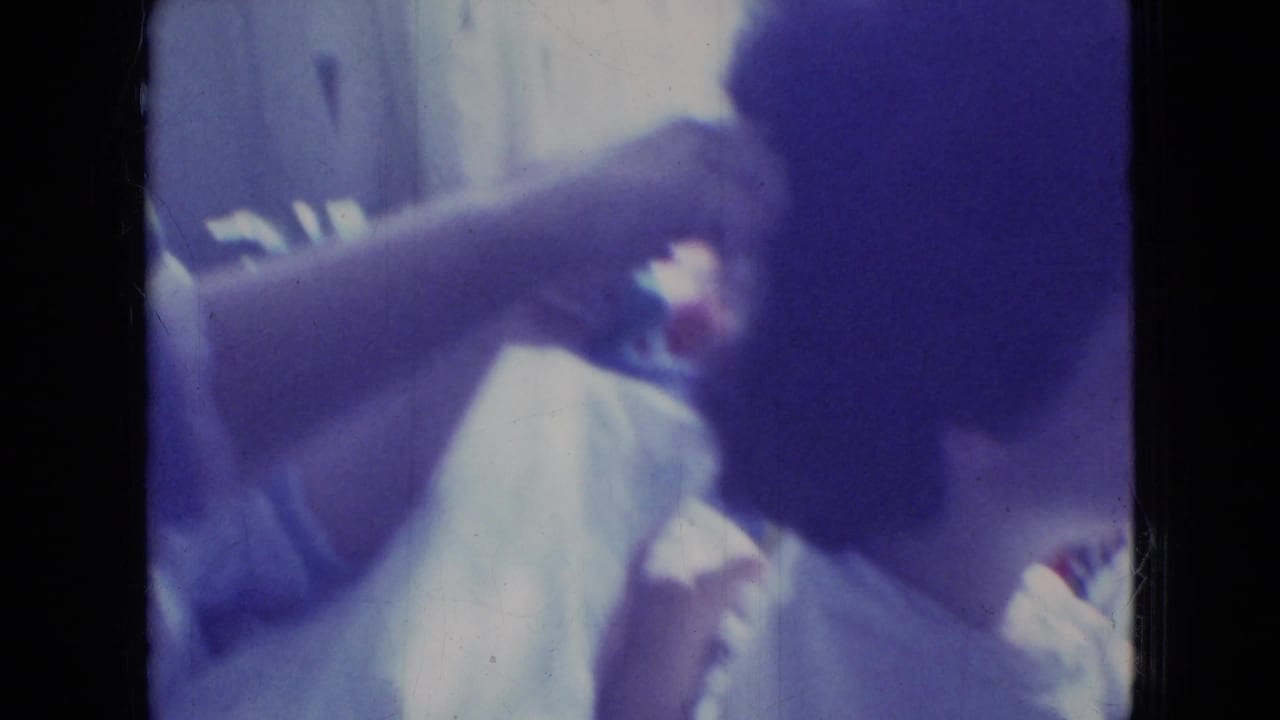 In a 1965 Wellsburg, Iowa 8mm home-movie frame, one person reaches in to comb or arrange another person's dark hair in a soft, color-shifted interior.