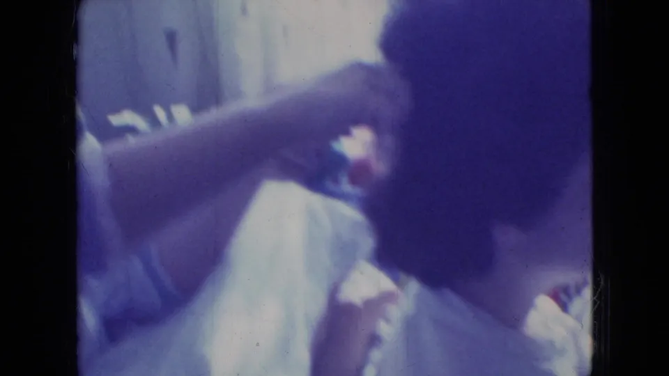 In a 1965 Wellsburg, Iowa 8mm home-movie frame, one person reaches in to comb or arrange another person's dark hair in a soft, color-shifted interior.