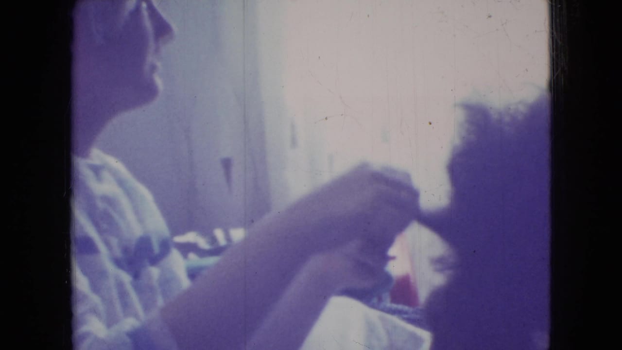 Seated adult reaches toward a dark-haired person during hair grooming.