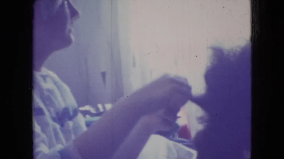 A seated adult in a light top appears to comb or style a dark-haired person's hair inside a bright private room, with 8mm color fading and frame borders visible.