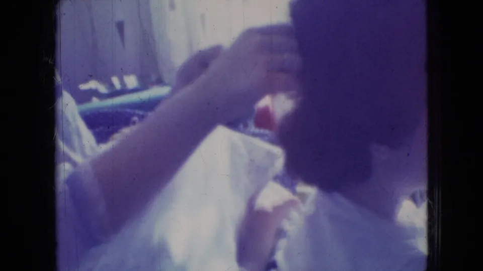 A close 8mm frame shows a hand working through a dark-haired person's hair in an intimate indoor grooming moment, with a light garment and faded purple color cast.
